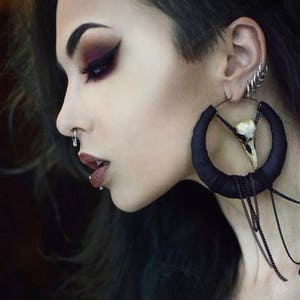 May include: A woman with dark hair and dark eye makeup is wearing a large black hoop earring with a white skull charm and a chain dangling from it. She is also wearing multiple smaller earrings in her ear.