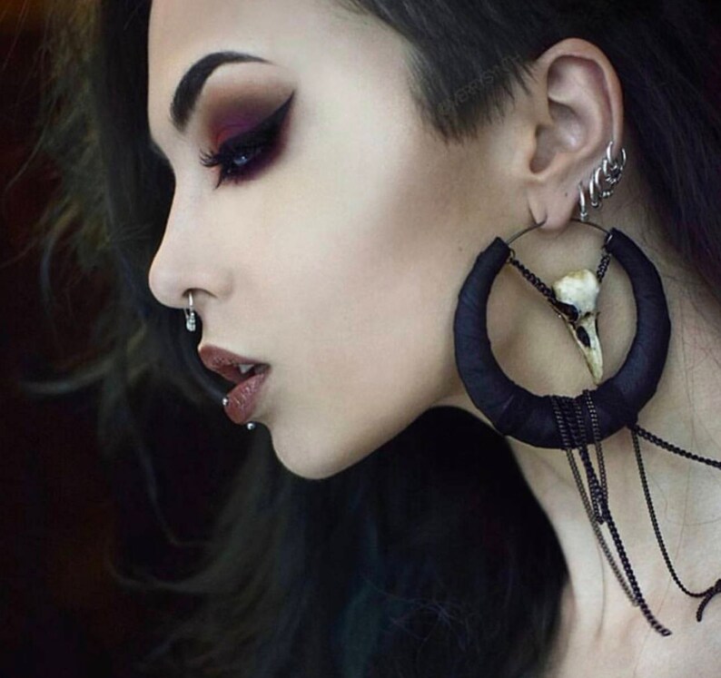 May include: A woman with dark hair and dark eye makeup is wearing a large black hoop earring with a white skull charm and a chain dangling from it. She is also wearing multiple smaller earrings in her ear.