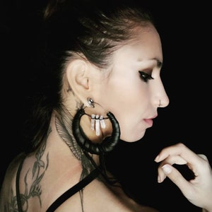 May include: A woman with a black leather hoop earring with white crystal accents. The earring is hanging from her left ear. She has a tattoo on her neck and shoulder.