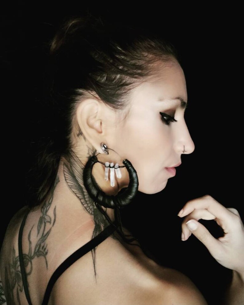 May include: A woman with a black leather hoop earring with white crystal accents. The earring is hanging from her left ear. She has a tattoo on her neck and shoulder.