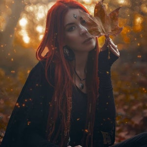 May include: A woman with long red hair is wearing a black top and a necklace with a pendant. She is holding a brown leaf in front of her face. The background is blurred and has a golden hue.