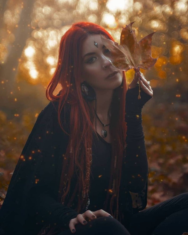 May include: A woman with long red hair is wearing a black top and a necklace with a pendant. She is holding a brown leaf in front of her face. The background is blurred and has a golden hue.