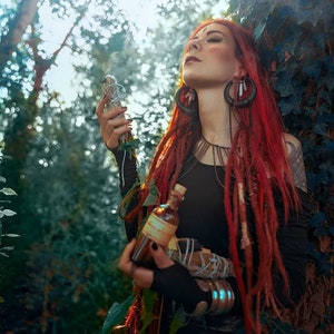 May include: A woman with long red dreadlocks stands in a forest, holding a smudge stick and a small bottle. She is wearing a black shirt and has a beaded necklace. The background is a lush green forest.