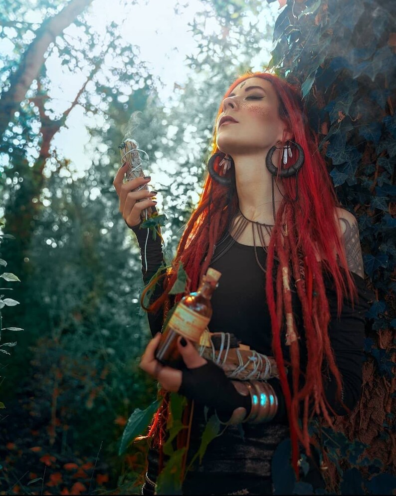 May include: A woman with long red dreadlocks stands in a forest, holding a smudge stick and a small bottle. She is wearing a black shirt and has a beaded necklace. The background is a lush green forest.