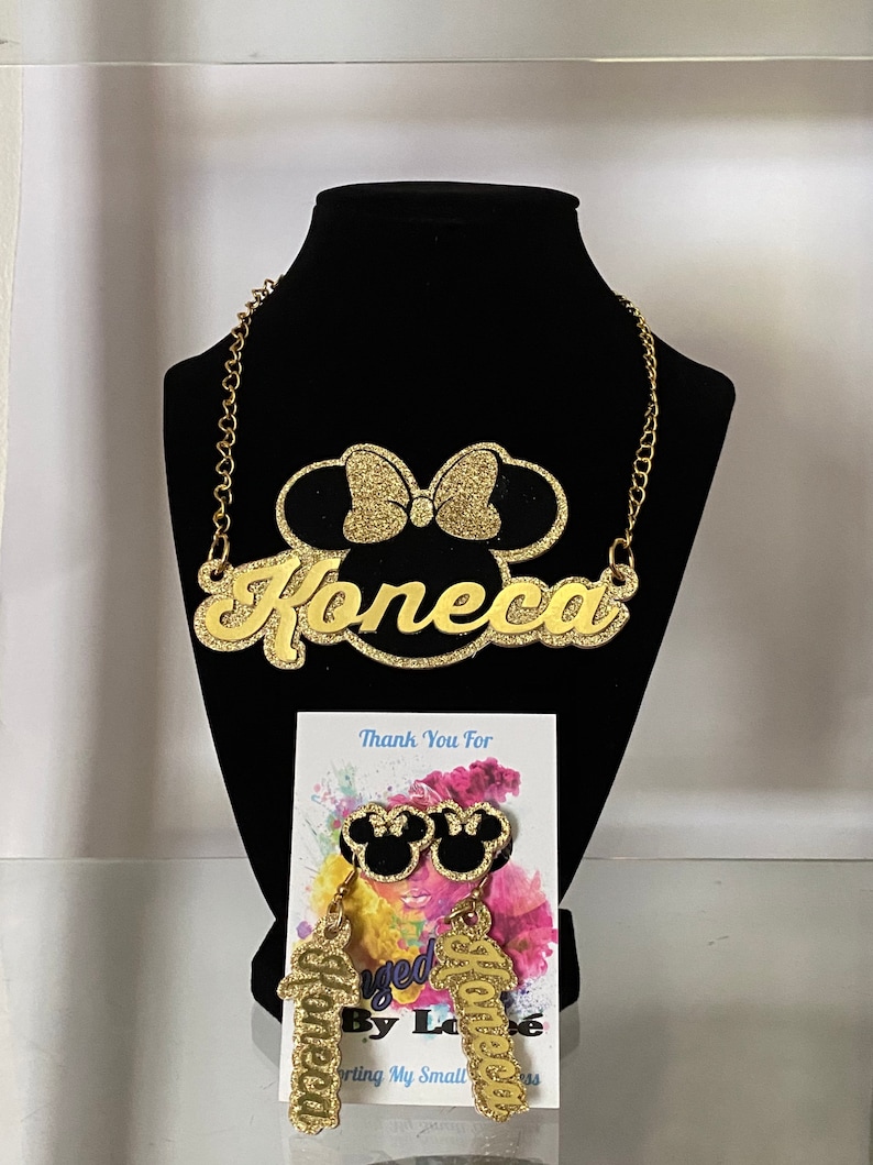 K&ouml;nnte beinhalten: Eine goldene Glitzerkette mit einem Mickey-Maus-Kopf-Anh&auml;nger und einem Namensschild mit der Aufschrift "Joneeca". Passende goldene Glitzerohrringe mit Mickey-Maus-Kopf-Anh&auml;ngern und dem Namen "Joneeca", der an ihnen h&auml;ngt. Die Ohrringe und die Kette befinden sich auf einem schwarzen Samt-Hintergrund.