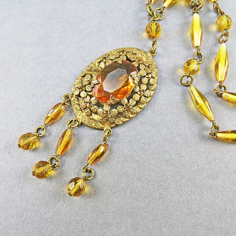 May include: A gold tone necklace with a large amber colored glass stone set in a filigree pendant. The necklace has a long chain with multiple strands of amber colored glass beads.