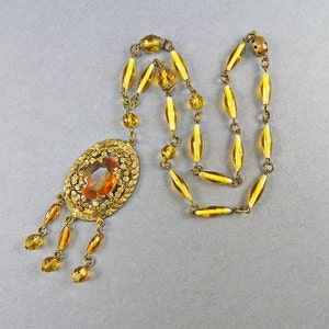 May include: A gold necklace with a large amber-colored glass pendant. The pendant is surrounded by a filigree design and has a teardrop shape. The necklace is made of small amber-colored glass beads.