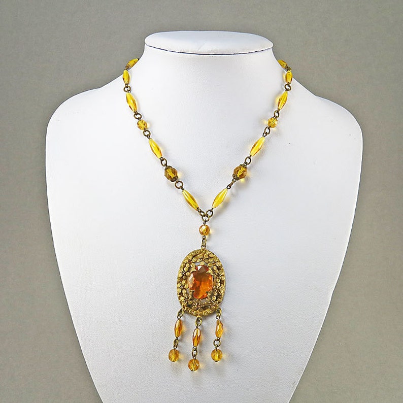 May include: A gold necklace with a large amber pendant and a chain of amber beads. The pendant has a detailed design and a large amber stone in the center.