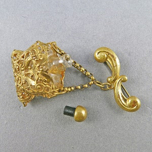 May include: A gold-tone filigree perfume bottle with a clear glass bottle and a chain attached to a gold-tone pin. The bottle has a separate gold-tone stopper.