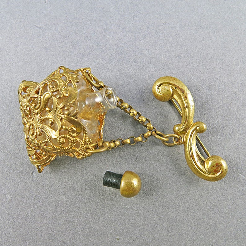 May include: A gold-tone filigree perfume bottle with a clear glass bottle and a chain attached to a gold-tone pin. The bottle has a separate gold-tone stopper.