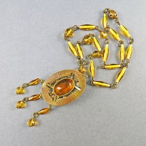 May include: A gold tone necklace with a large oval pendant featuring a faceted amber stone. The pendant has a geometric design and hangs from a chain of amber glass beads.
