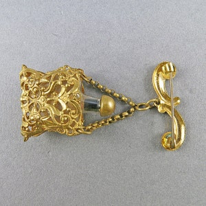 May include: Gold filigree perfume bottle with a chain and a pin back. The bottle is empty and has a clear glass interior.