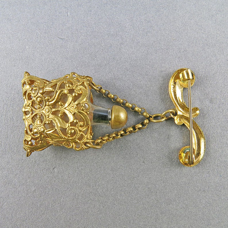 May include: Gold filigree perfume bottle with a chain and a pin back. The bottle is empty and has a clear glass interior.