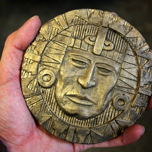 Puede incluir: Una pieza decorativa circular dorada que presenta un rostro detallado con un tocado. El rostro tiene los ojos cerrados y una expresión serena. La pieza se sostiene en una mano, mostrando su tamaño y textura.