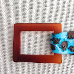 May include: A square, tortoiseshell-colored buckle with a clear center is shown. A section of blue fabric with a brown animal print pattern is attached to the buckle. The fabric is stitched with blue thread.