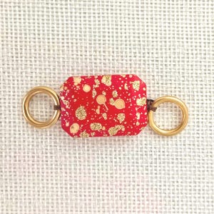 May include: A rectangular red bead with gold flecks and two gold rings attached with brown cord. The bead is approximately 1 inch long and 0.5 inches wide. The gold rings are about 0.75 inches in diameter. The item is on a white woven background.
