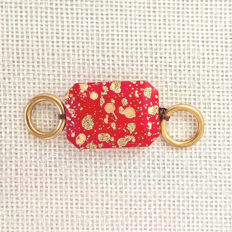 May include: A rectangular red bead with gold flecks and two gold rings attached with brown cord. The bead is approximately 1 inch long and 0.5 inches wide. The gold rings are about 0.75 inches in diameter. The item is on a white woven background.