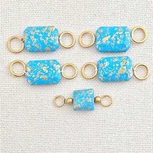 May include: Several turquoise and gold-flecked rectangular beads with gold-colored connecting rings. The beads are arranged in a pattern on a light-colored surface. These beads are likely for jewelry making.