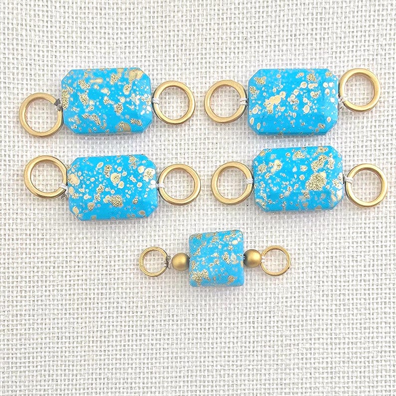 May include: Several turquoise and gold-flecked rectangular beads with gold-colored connecting rings. The beads are arranged in a pattern on a light-colored surface. These beads are likely for jewelry making.