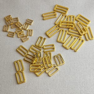 Puede incluir: Una colección de ajustadores y deslizadores de tirantes de sujetador de metal dorado. Los ajustadores son rectangulares con una barra central, mientras que los deslizadores tienen un diseño de doble bucle. Estos hallazgos están dispersos sobre una tela blanca.