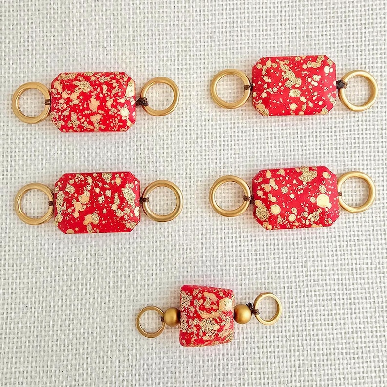May include: Five red rectangular beads with gold splatter and gold-toned rings. The beads are connected by small gold beads and dark brown string. The beads are displayed on a white textured surface.