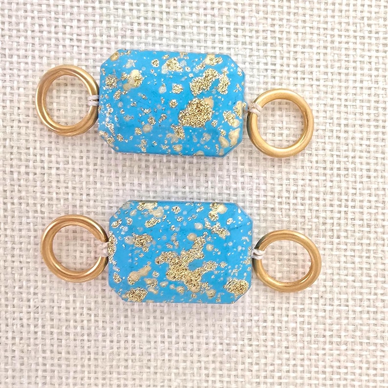 May include: Two rectangular turquoise beads with gold flecks and gold-colored ring connectors. The beads are a vibrant blue with irregular gold patterns. The connectors are attached with a small, off-white string.