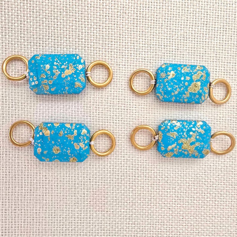 May include: Four turquoise and gold rectangular jewelry clasps. Each clasp has a rectangular turquoise stone with gold flecks, connected to two gold-colored rings. The clasps are arranged on a textured, off-white surface.