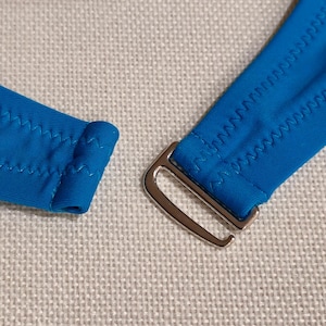 May include: Close-up of a bright blue bikini top strap with a silver metal clasp. The strap is sewn with a zig-zag stitch.