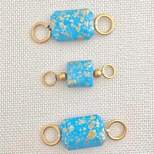 May include: Three turquoise-colored rectangular beads with gold flecks and gold-colored ring connectors. The beads are arranged vertically against a neutral background, showcasing their design and color variations. These beads are suitable for jewelry making.