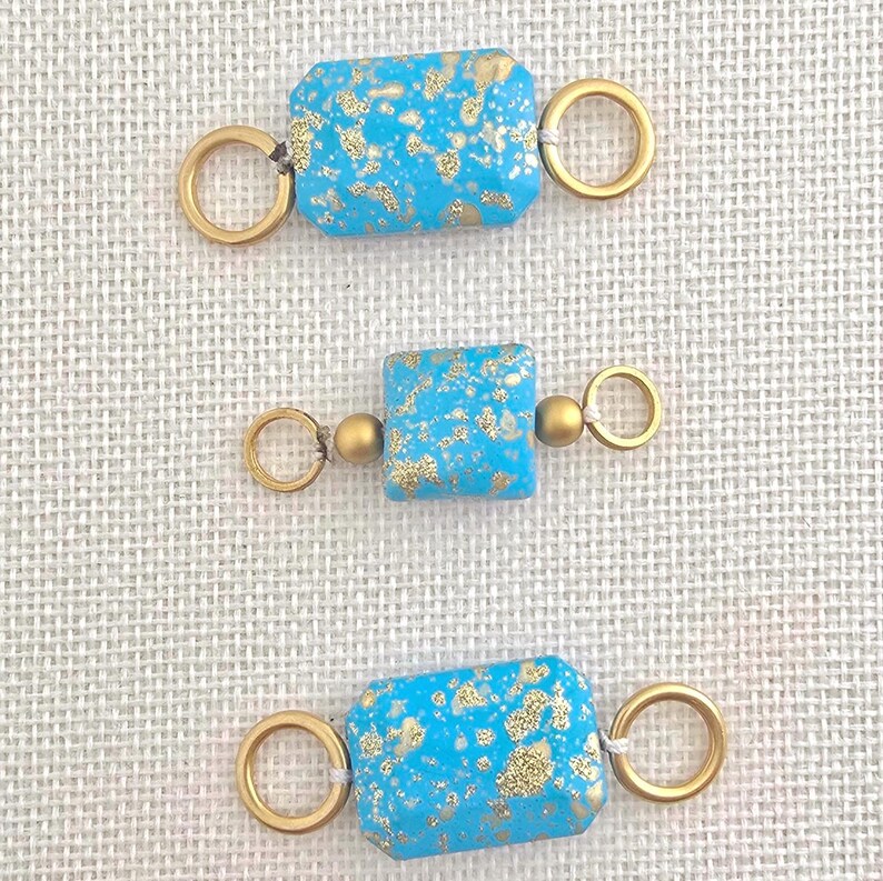 May include: Three turquoise-colored rectangular beads with gold flecks and gold-colored ring connectors. The beads are arranged vertically against a neutral background, showcasing their design and color variations. These beads are suitable for jewelry making.