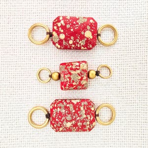 May include: Three handmade jewelry components. Each features a red, rectangular or square bead with gold speckles, connected to gold-colored rings and small beads with dark brown cord. The components are displayed on a white textured surface.