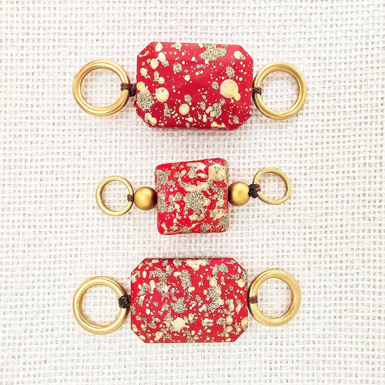 May include: Three handmade jewelry components. Each features a red, rectangular or square bead with gold speckles, connected to gold-colored rings and small beads with dark brown cord. The components are displayed on a white textured surface.