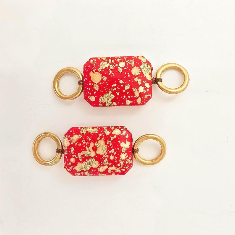 May include: Two rectangular red fabric pieces with gold splatter designs, connected to gold-colored rings on each end. The rings are attached with brown cords. The items are displayed on a white background.