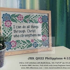 May include: Cross-stitch framed artwork with the text "I can do all things through Christ who strengthens me." The design features floral and leaf patterns in blue, pink, and green. The frame is light wood. A potted plant is in the foreground.