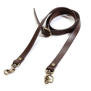 May include: A dark brown leather bag strap with brass-colored swivel clasps and a buckle. The strap is long and thin, designed to be adjustable. The hardware is a warm, antique brass color.