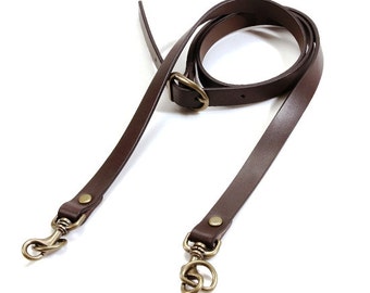 May include: A dark brown leather bag strap with brass-colored swivel clasps and a buckle. The strap is long and thin, designed to be adjustable. The hardware is a warm, antique brass color.