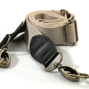 May include: A beige canvas camera strap with black leather accents and antique gold metal clasps.