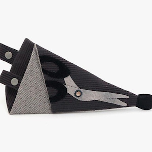 May include: A black and gray fabric scissor holder with a gray and white patterned fabric on one side. The holder has a black button on the top and a black button on the side. The holder is shaped like a triangle and has a black pom-pom on the end.
