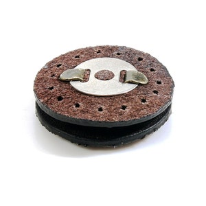 May include: A brown leather sanding disc with a metal center and a metal clip. The disc has a rough texture and is designed for sanding wood.