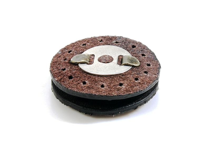 May include: A brown leather sanding disc with a metal center and a metal clip. The disc has a rough texture and is designed for sanding wood.