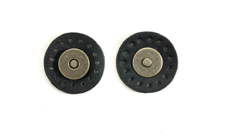 May include: Two black leather magnetic closures with metal accents. The closures are round and have a circle of small holes around the edge. One closure has the text "SAFE LOCK" printed on the metal accent.