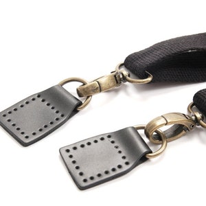 May include: Two black leather straps with brass hardware and metal clasps. The straps have a square shape with a stitched border.
