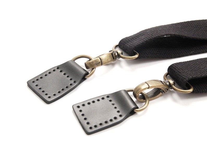 May include: Two black leather straps with brass hardware and metal clasps. The straps have a square shape with a stitched border.