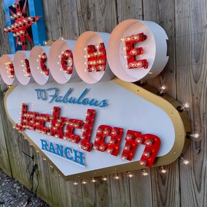 K&ouml;nnte beinhalten: Ein wei&szlig;es, goldenes und rotes Schild mit Gl&uuml;hbirnen, auf dem "Willkommen auf der Fabulous Jackolan Ranch" steht.
