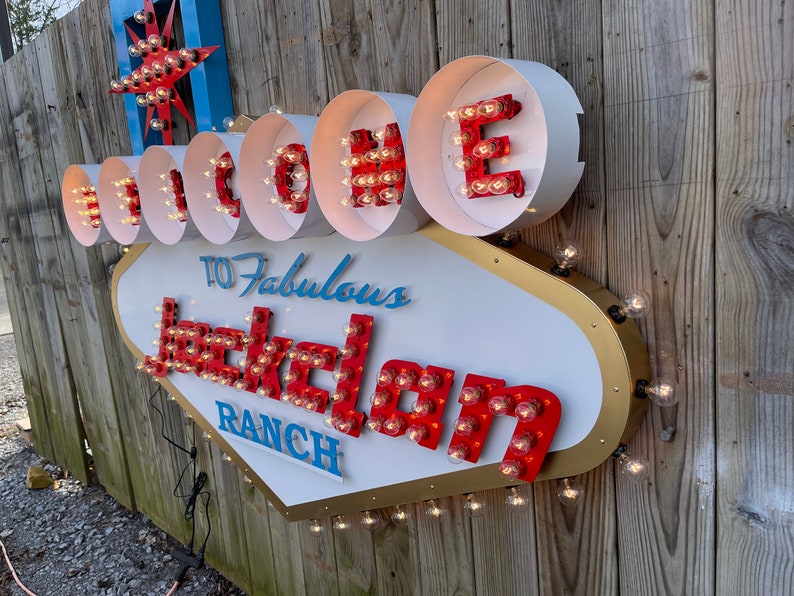 K&ouml;nnte beinhalten: Ein wei&szlig;es, goldenes und rotes Schild mit Gl&uuml;hbirnen, auf dem "Willkommen auf der Fabulous Jackolan Ranch" steht.