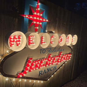K&ouml;nnte beinhalten: Ein beleuchtetes Schild in Wei&szlig; und Rot mit der Aufschrift "Welcome to Fabulous Jackson Ranch". Das Schild ist aus Metall und hat eine Diamantform. Die Buchstaben sind rot umrandet und haben wei&szlig;e Gl&uuml;hbirnen im Inneren. Das Schild ist an einer Holzwand befestigt.