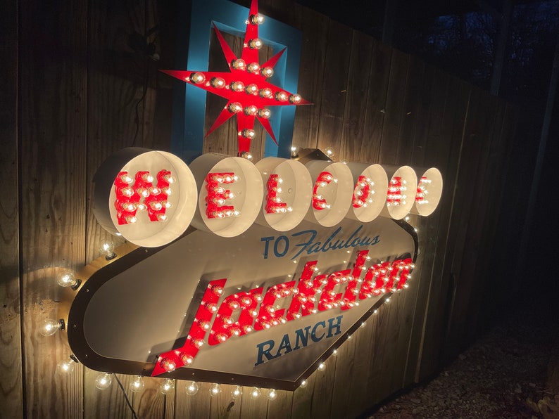 K&ouml;nnte beinhalten: Ein beleuchtetes Schild in Wei&szlig; und Rot mit der Aufschrift "Welcome to Fabulous Jackson Ranch". Das Schild ist aus Metall und hat eine Diamantform. Die Buchstaben sind rot umrandet und haben wei&szlig;e Gl&uuml;hbirnen im Inneren. Das Schild ist an einer Holzwand befestigt.