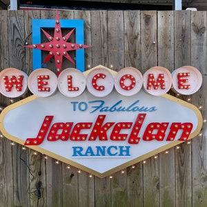 K&ouml;nnte beinhalten: Ein wei&szlig;es und goldenes Schild mit roten Gl&uuml;hbirnen, die "WELCOME TO Fabulous Jackelan RANCH" buchstabieren. Das Schild hat die Form eines Diamanten und einen goldenen Rand. Das Schild h&auml;ngt an einem Holzzaun.