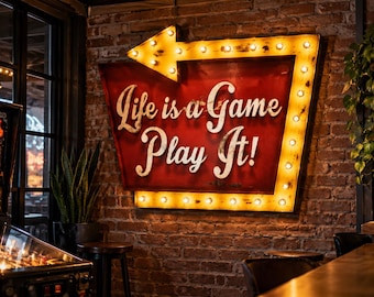 Könnte beinhalten: Ein großes, pfeilförmiges Wandschild mit rotem Hintergrund und weißer Aufschrift "Life is a Game Play It!". Das Schild ist von einer Reihe leuchtender gelber Glühbirnen umrahmt und an einer rustikalen Wand aus rotem Backstein befestigt.