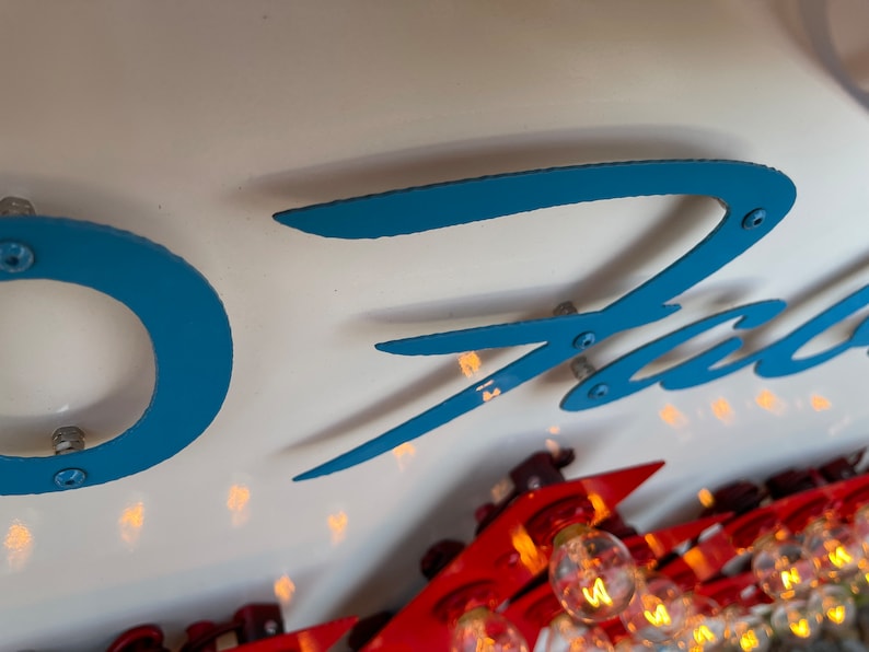 K&ouml;nnte beinhalten: Nahaufnahme eines blauen und wei&szlig;en Schilds mit dem Wort "Fast" in einer stilisierten Schrift. Das Schild hat mehrere kleine, runde, silberne Lichter an den R&auml;ndern.
